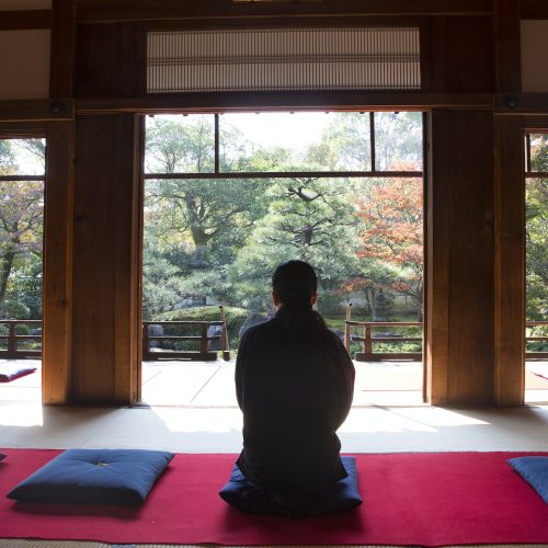 Rear view of man sitting on blue cushion on floor in traditional Japanese building.